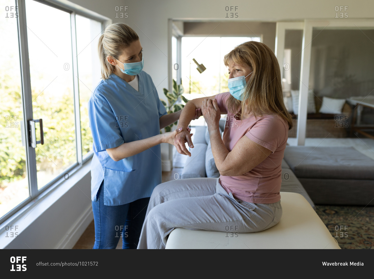 Senior Caucasian woman at home visited by Caucasian female nurse, stretching her arm, wearing face masks. Medical care at home during Covid 19 Coronavirus quarantine.