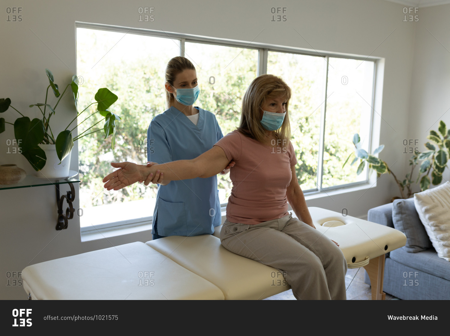 Senior Caucasian woman at home visited by Caucasian female nurse, stretching her arm, wearing face masks. Medical care at home during Covid 19 Coronavirus quarantine.