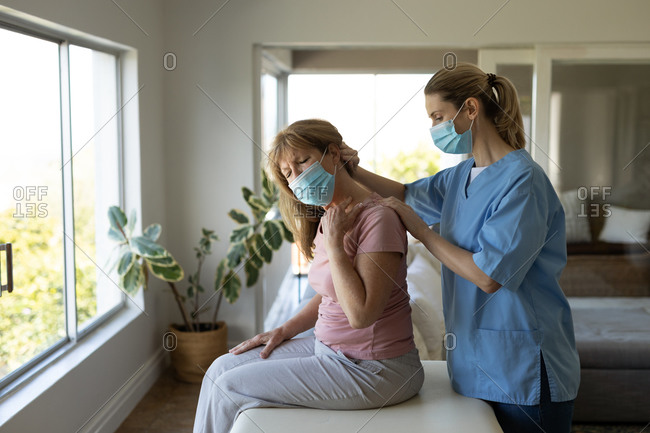 Senior Caucasian woman at home visited by Caucasian female nurse, stretching her neck, wearing face masks. Medical care at home during Covid 19 Coronavirus quarantine.