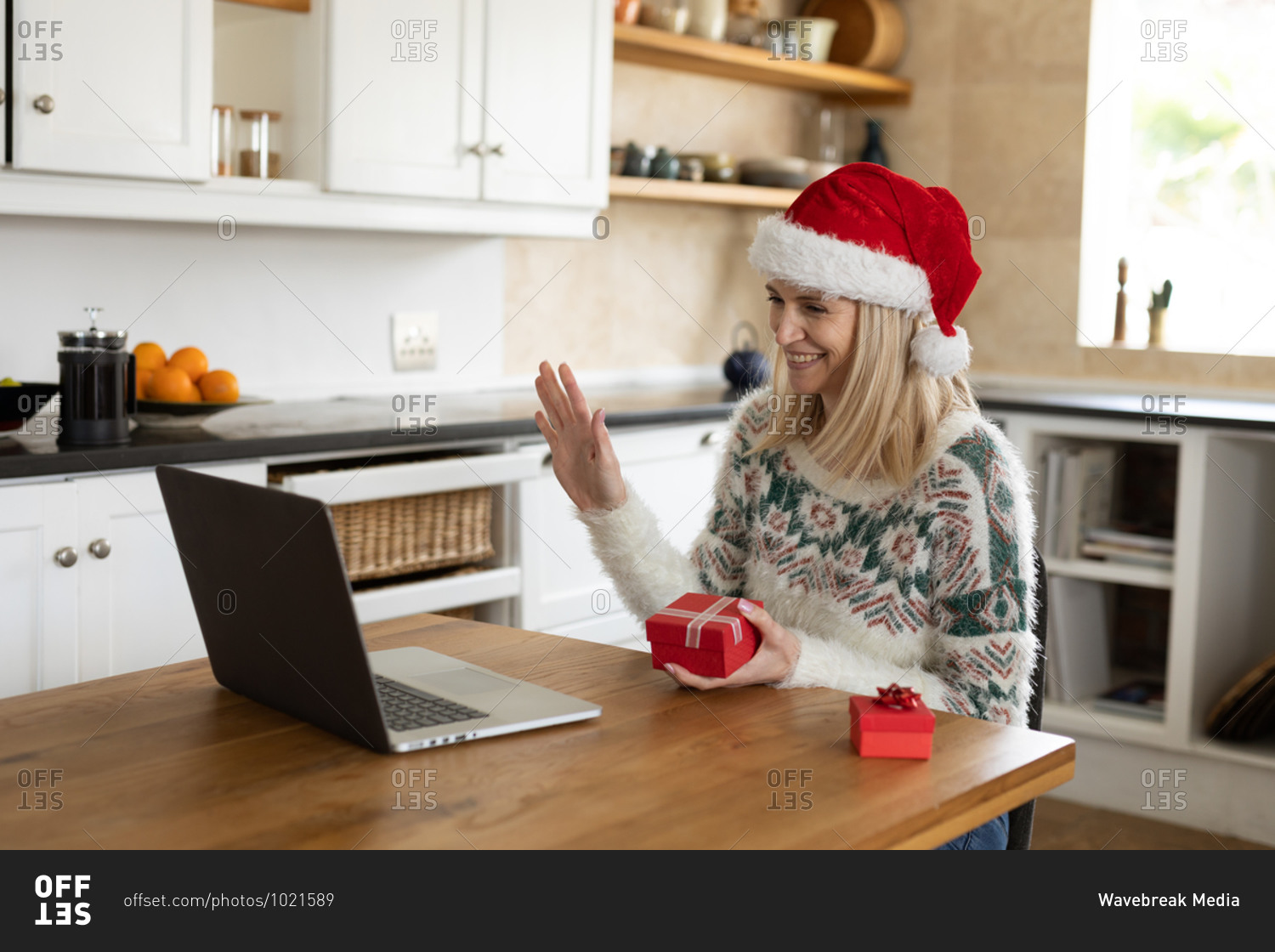 Caucasian woman spending time at home, sitting in kitchen at Christmas wearing Santa hat, using laptop with presents on table. Social distancing during Covid 19 Coronavirus quarantine.