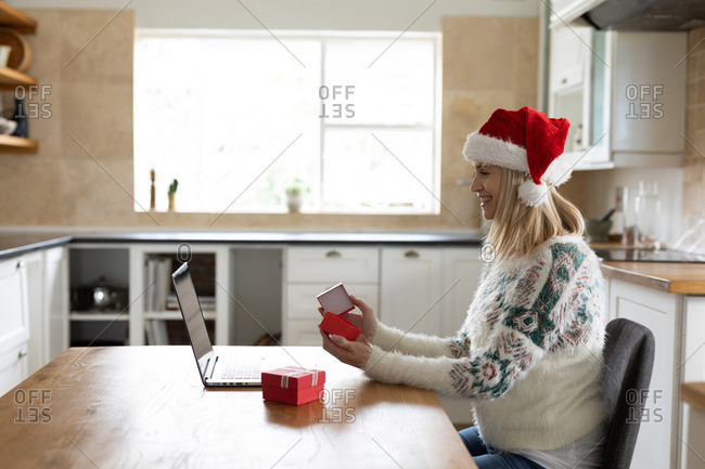 Caucasian woman spending time at home, sitting in kitchen at Christmas wearing Santa hat, using laptop with presents on table. Social distancing during Covid 19 Coronavirus quarantine.