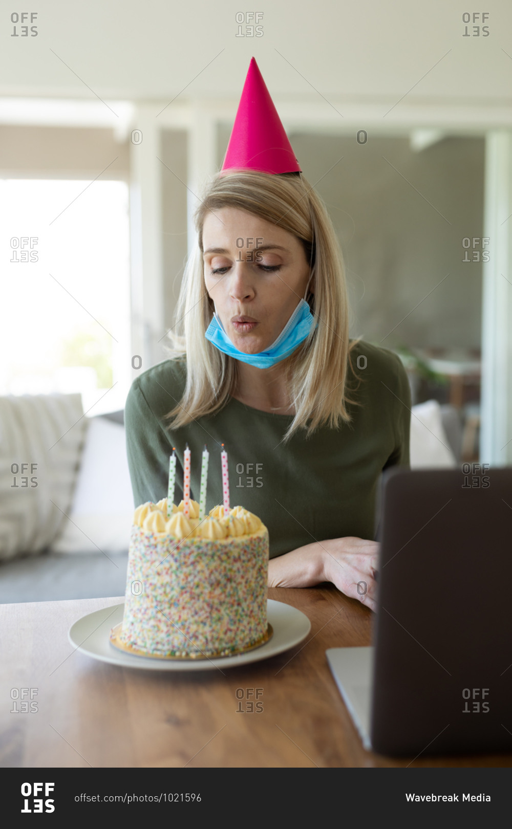 Caucasian woman spending time at home, sitting in living room with birthday cake and using laptop, blowing candles. Social distancing during Covid 19 Coronavirus quarantine.