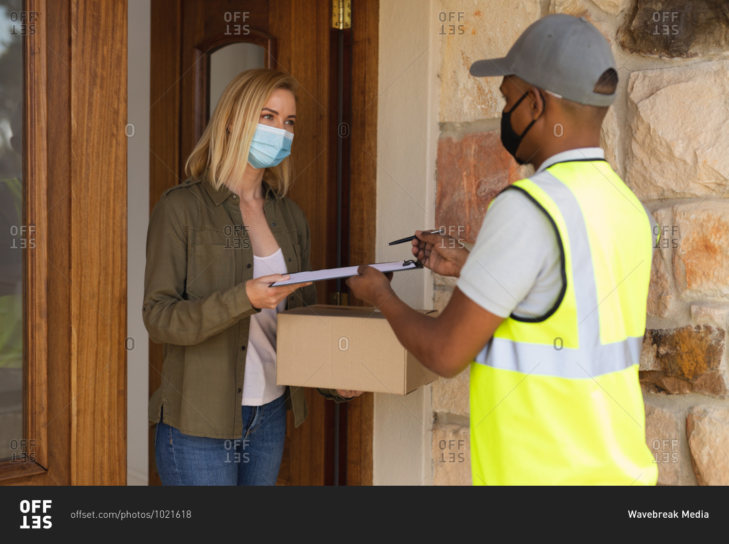 Caucasian woman spending time at home, wearing face mask, receiving a package from delivery man. Social distancing during Covid 19 Coronavirus quarantine lockdown.