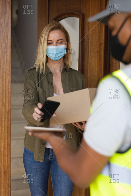 Caucasian woman spending time at home, wearing face mask, receiving a package from delivery man and paying by smartphone. Social distancing during Covid 19 Coronavirus quarantine lockdown.