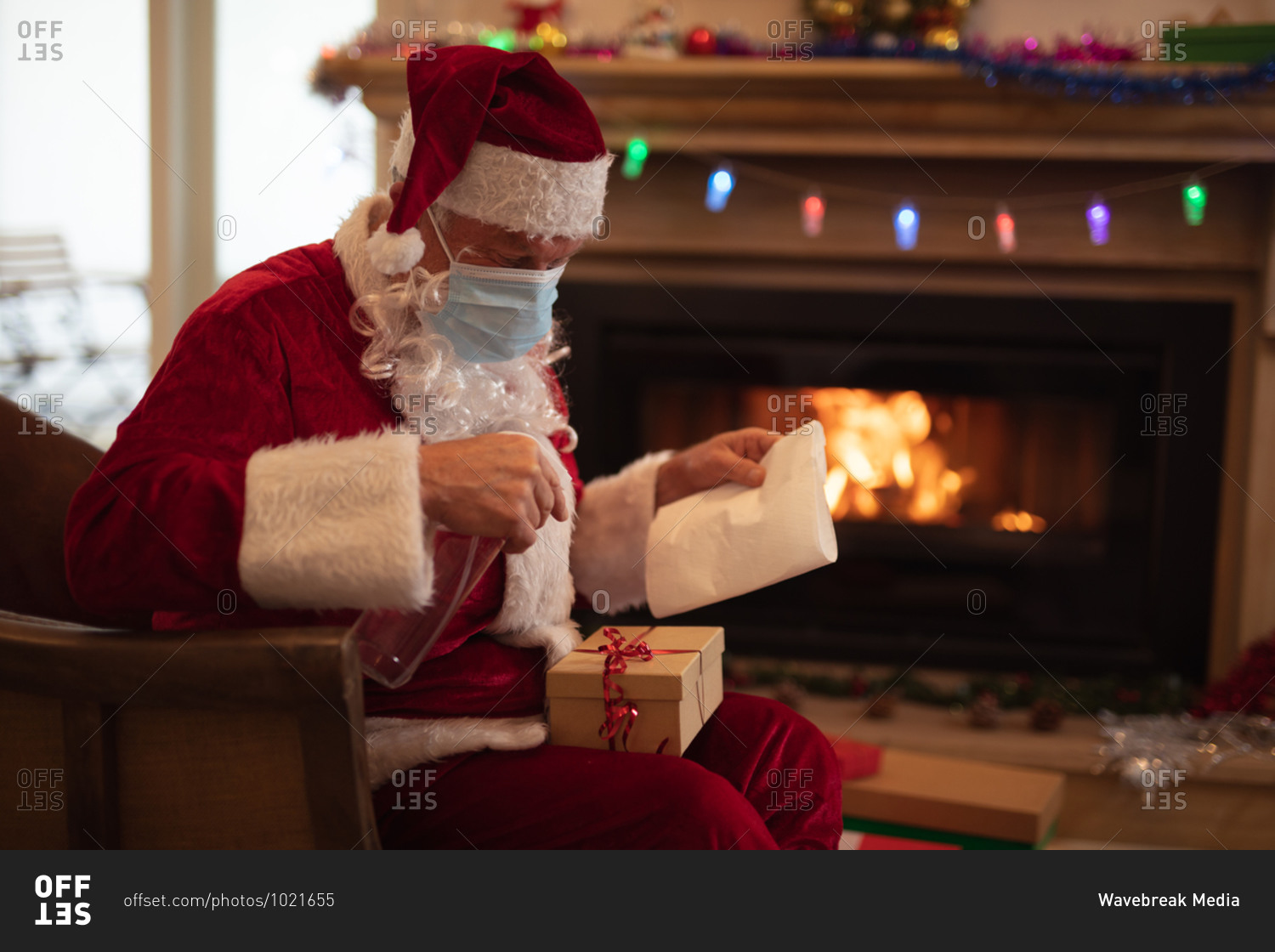 Senior Caucasian man at home dressed as Father Christmas, wearing face mask, disinfecting a present. Social distancing during Covid 19 Coronavirus quarantine lockdown.