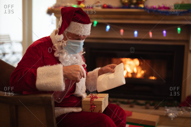 Senior Caucasian man at home dressed as Father Christmas, wearing face mask, disinfecting a present. Social distancing during Covid 19 Coronavirus quarantine lockdown.