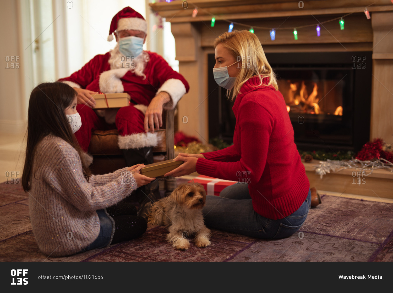Senior Caucasian man, his adult daughter and granddaughter at home dressed as Father Christmas, wearing face mask, giving presents. Social distancing during Covid 19 Coronavirus quarantine lockdown.