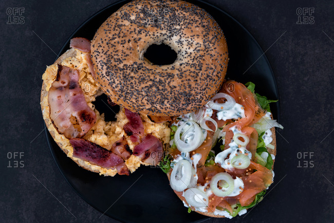 Two different bagels with lox and bacon on a black plate in dark setting