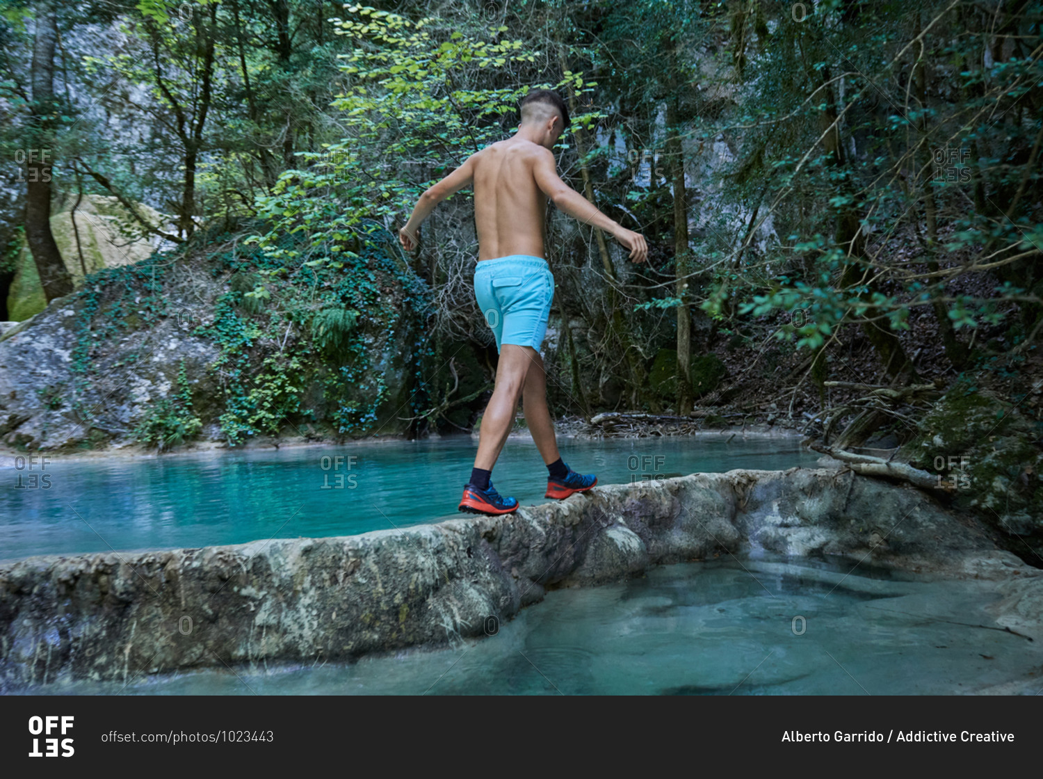 Traveling young man in swimwear crossing river with turquoise water in mountainous area while enjoying summer vacation