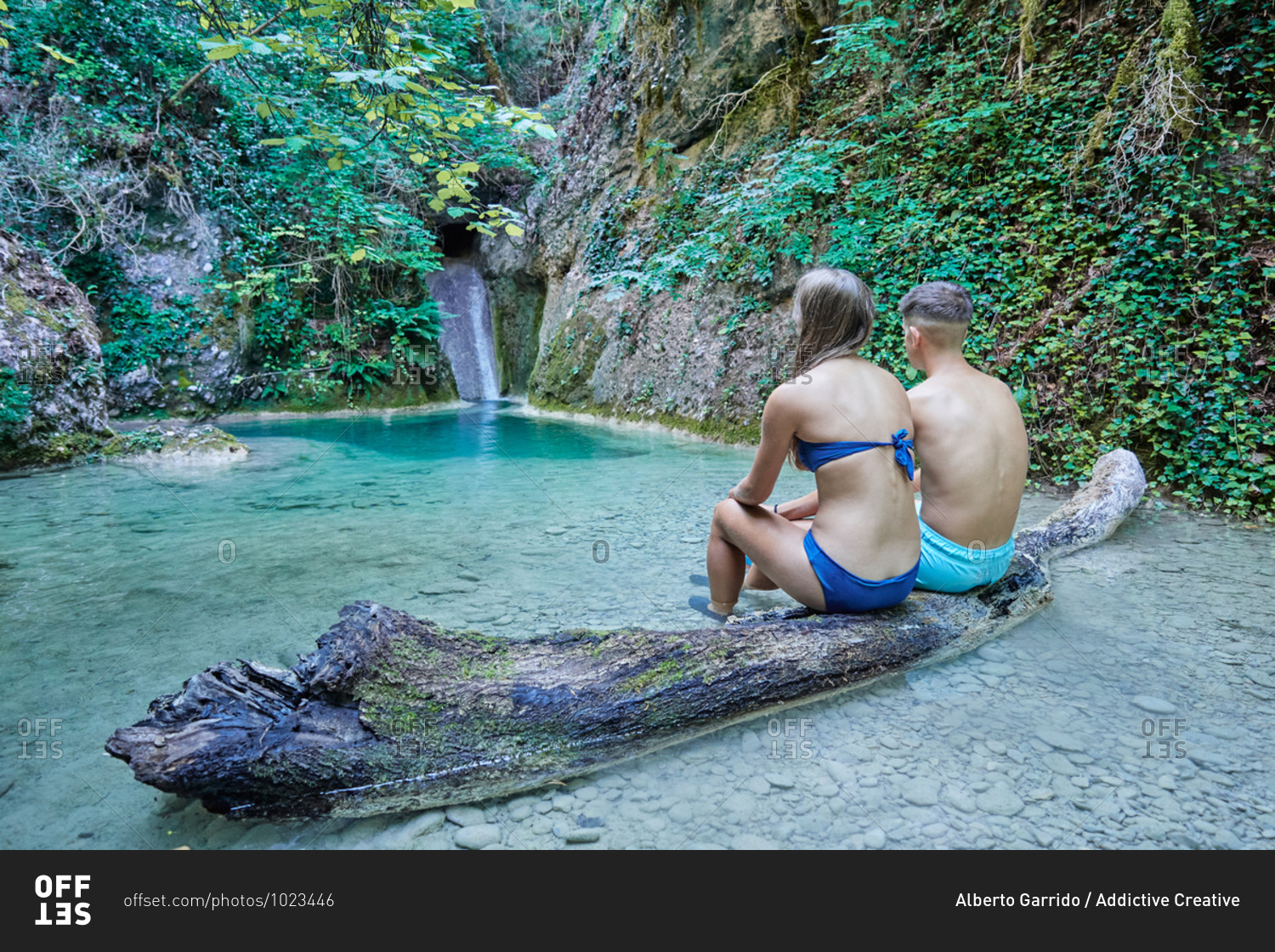 Back view of anonymous couple of tourists wearing bikini and shorts sitting on log in clear water of river in mountains and admiring amazing landscape