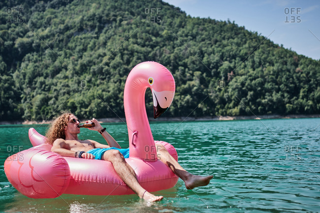 Relaxed male with bottle of refreshing drink lying on inflatable ring in shape of flamingo and floating on lake during summer vacation