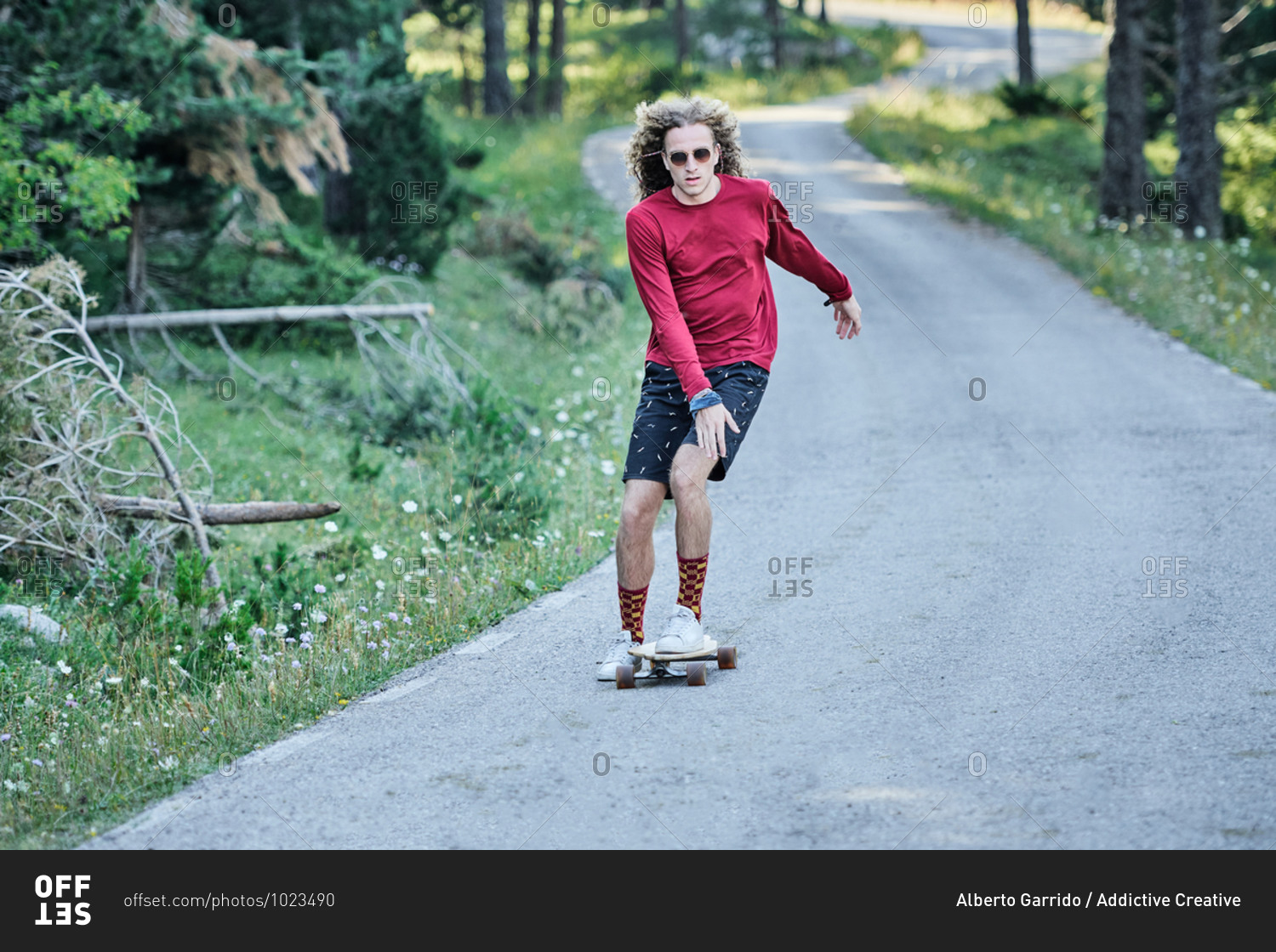 Concentrated male hipster in fancy clothes and with long hair riding skateboard along road in park in summer