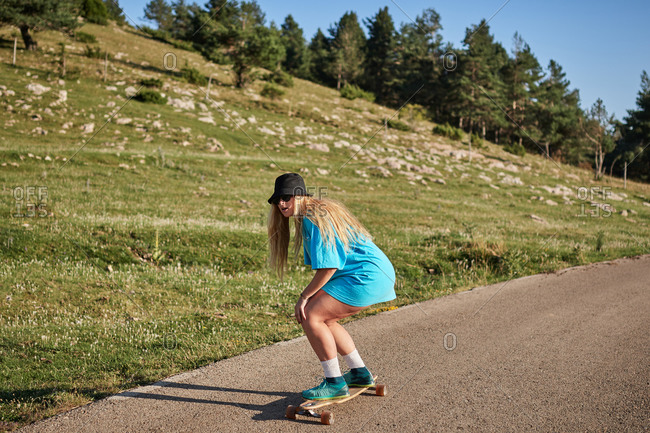 Full body of focused female skater riding skateboard along rural road while enjoying weekend in summer on background of mountains