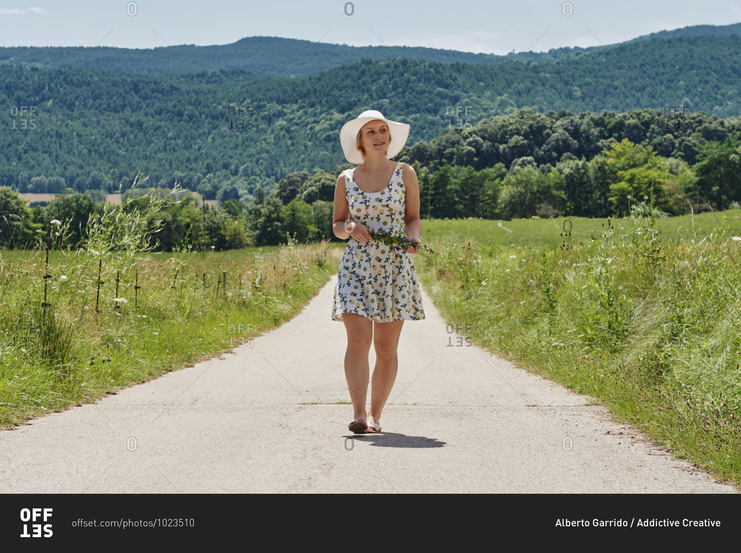 Delighted female wearing dress and sunhat walking with bunch of flowers along road on background of mountains and looking away