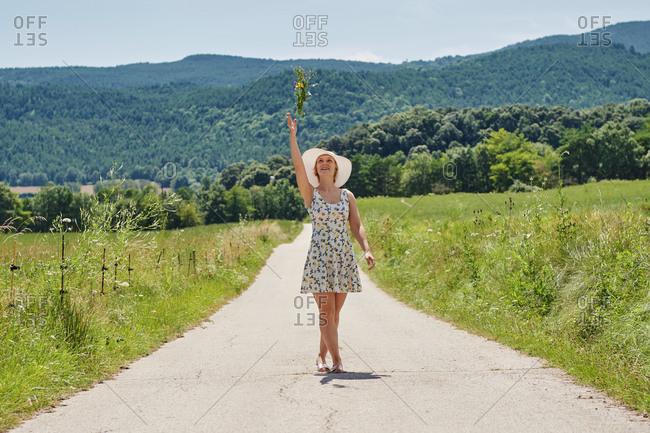 Delighted female wearing dress and sunhat walking with bunch of flowers along road on background of mountains and looking up