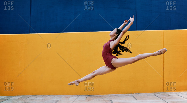 Side view of flexible ballerina wearing pointe shoes and bodysuit in moment of jumping with raised arms in city