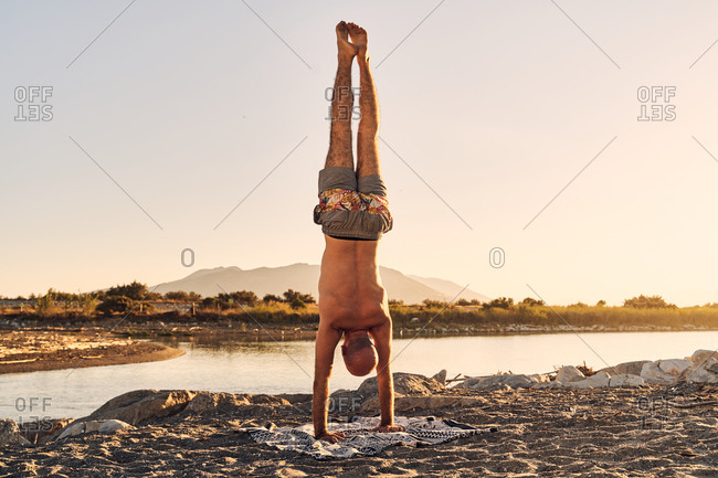 Back view of shirtless male balancing in Handstand pose while practicing yoga on river bank during amazing sundown
