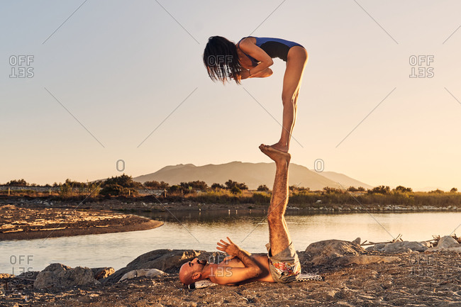 Side view of slim woman standing on feet of male lying on shore with Namaste hands while practicing mindfulness together