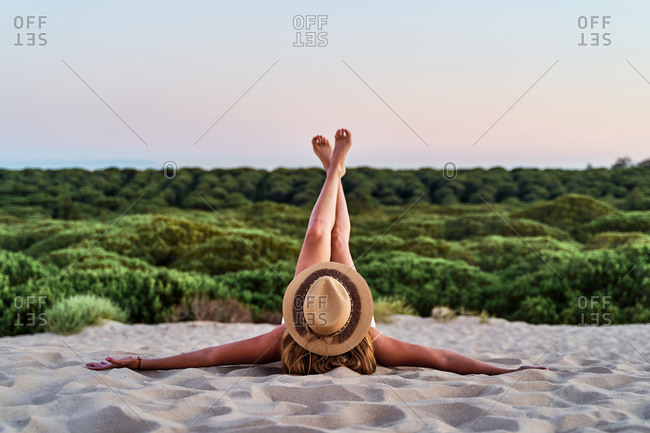 Anonymous female in straw hat relaxing on sandy shore with raised and crossed legs during sunset in summer