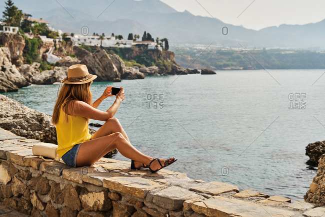 Unrecognizable woman sitting on stone border and taking photo of sea while resting on embankment on sunny day in Nerja, Spain