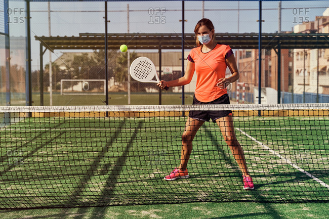 Focused sportswoman in activewear and mask with racket playing paddle tennis on sports ground in summer