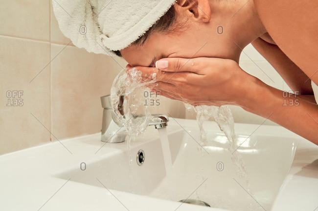 Side view of unrecognizable female in towel turban washing face with fresh water in bathroom at home