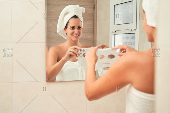 Focused female in towel turban applying moisturizing mask on face while standing in front of mirror after shower in bathroom