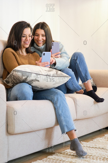 Positive female friends sitting on couch in cozy room and reading messages on social media on mobile phones while relaxing during weekend at home