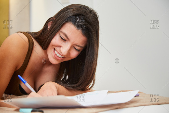 Cheerful female student lying on bed at home and writing on paper while preparing homework assignment