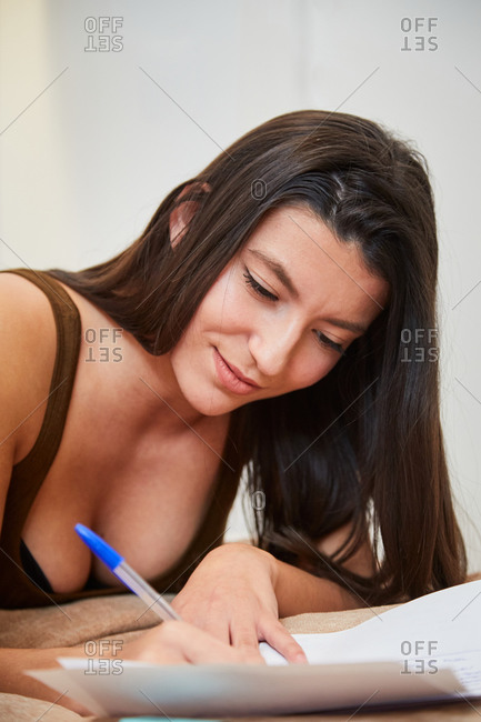 Cheerful female student lying on bed at home and writing on paper while preparing homework assignment