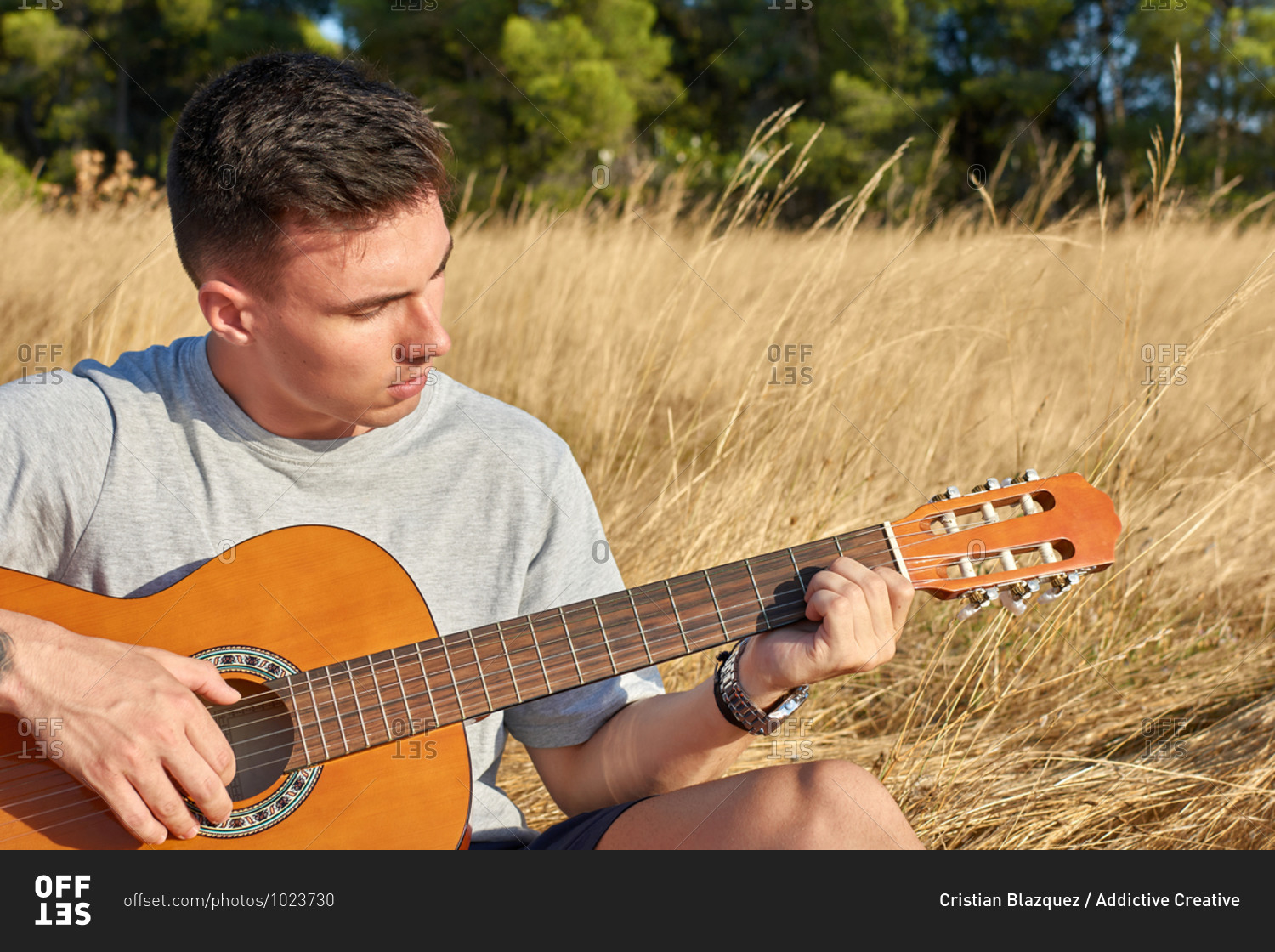 Young talented male guitarist sitting in dried meadow and playing acoustic musical instrument on sunny day