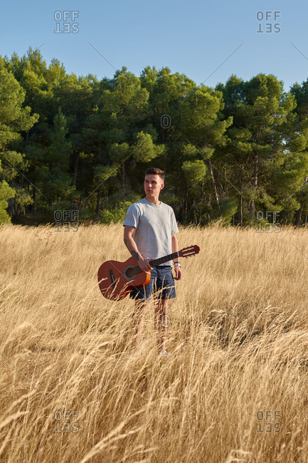 Peaceful male musician with guitar in case walking along dried field in summer and looking away