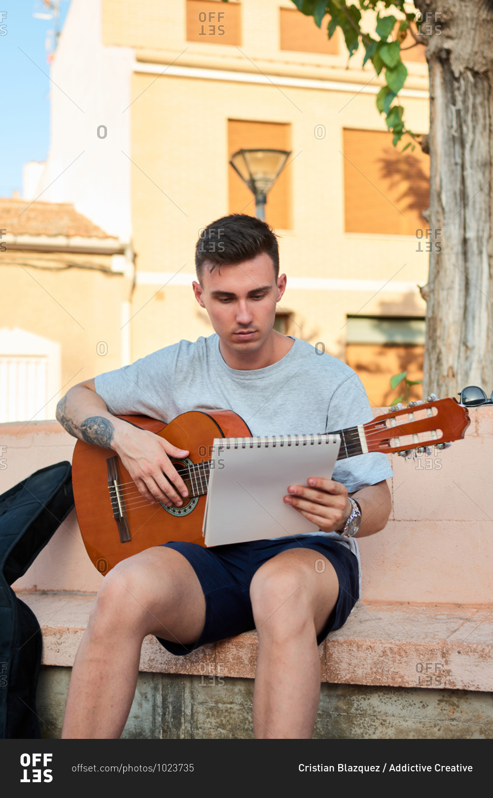 Creative male musician sitting on stone bench on street and reading notes while preparing for playing guitar