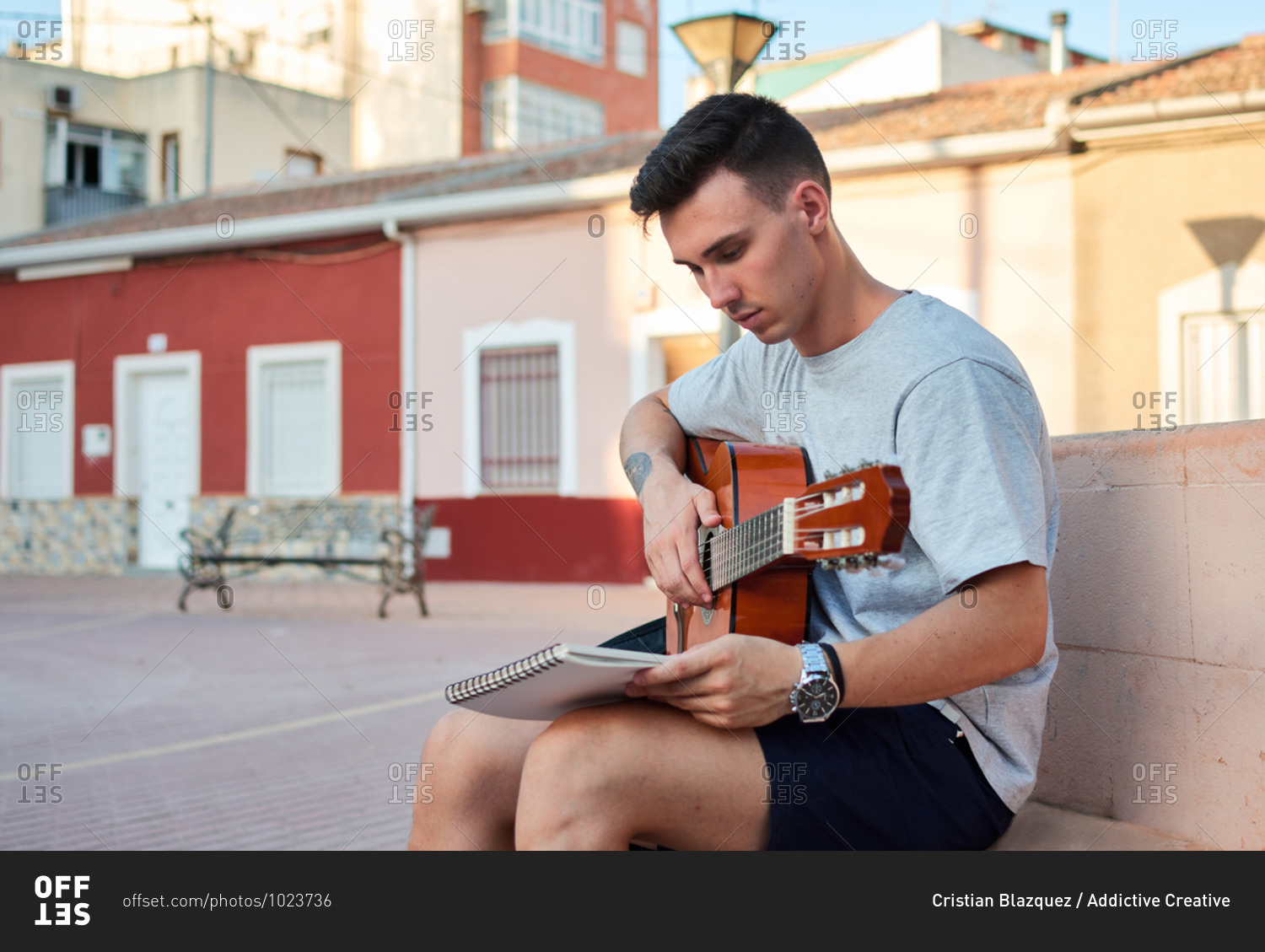 Creative male musician sitting on stone bench on street and reading notes while preparing for playing guitar