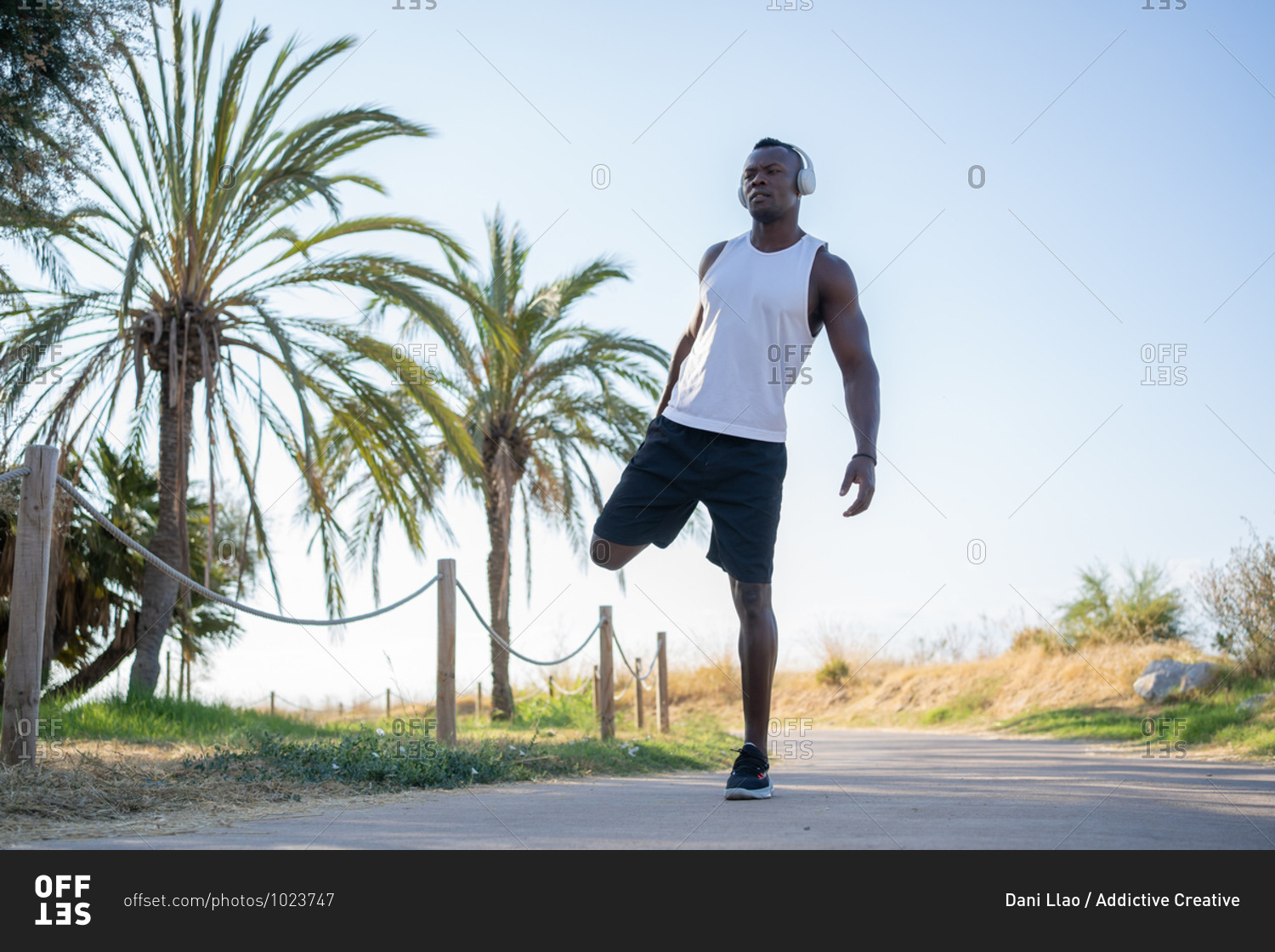 Full body of young muscular African American sportsman in headphones and sunglasses doing stretching exercise while preparing for jogging in city park in summer day