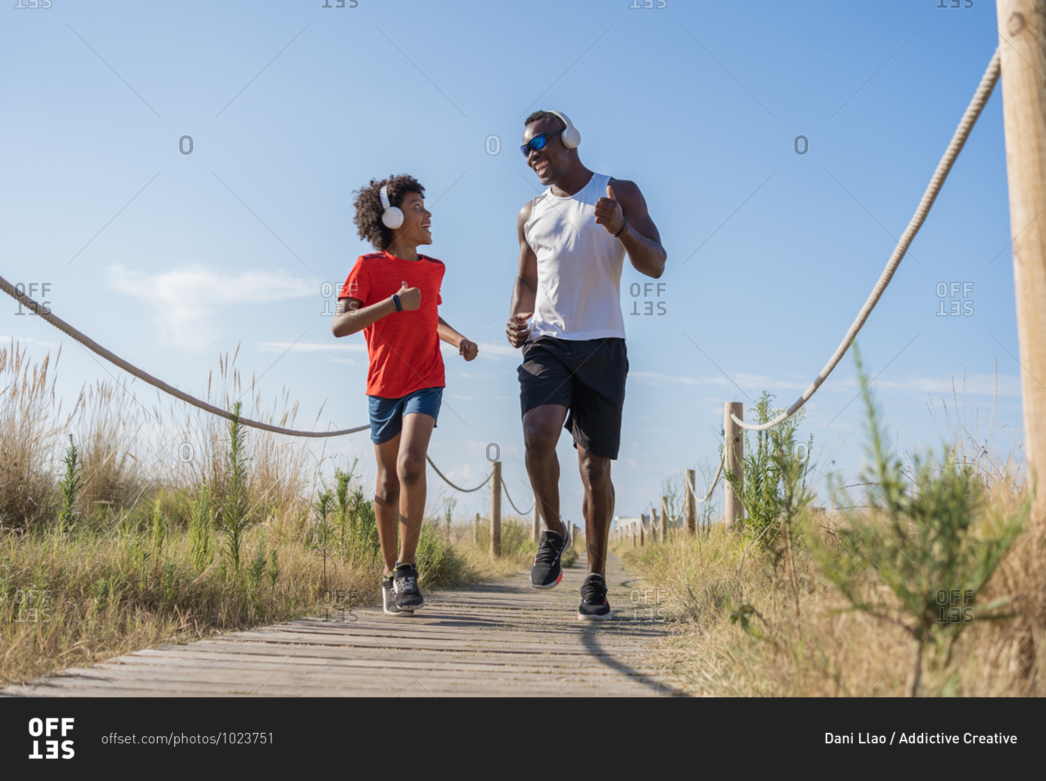 Low angle full body of cheerful active African American dad and son in sportswear and headphones jogging along fenced way while training together in sunny summer day in countryside