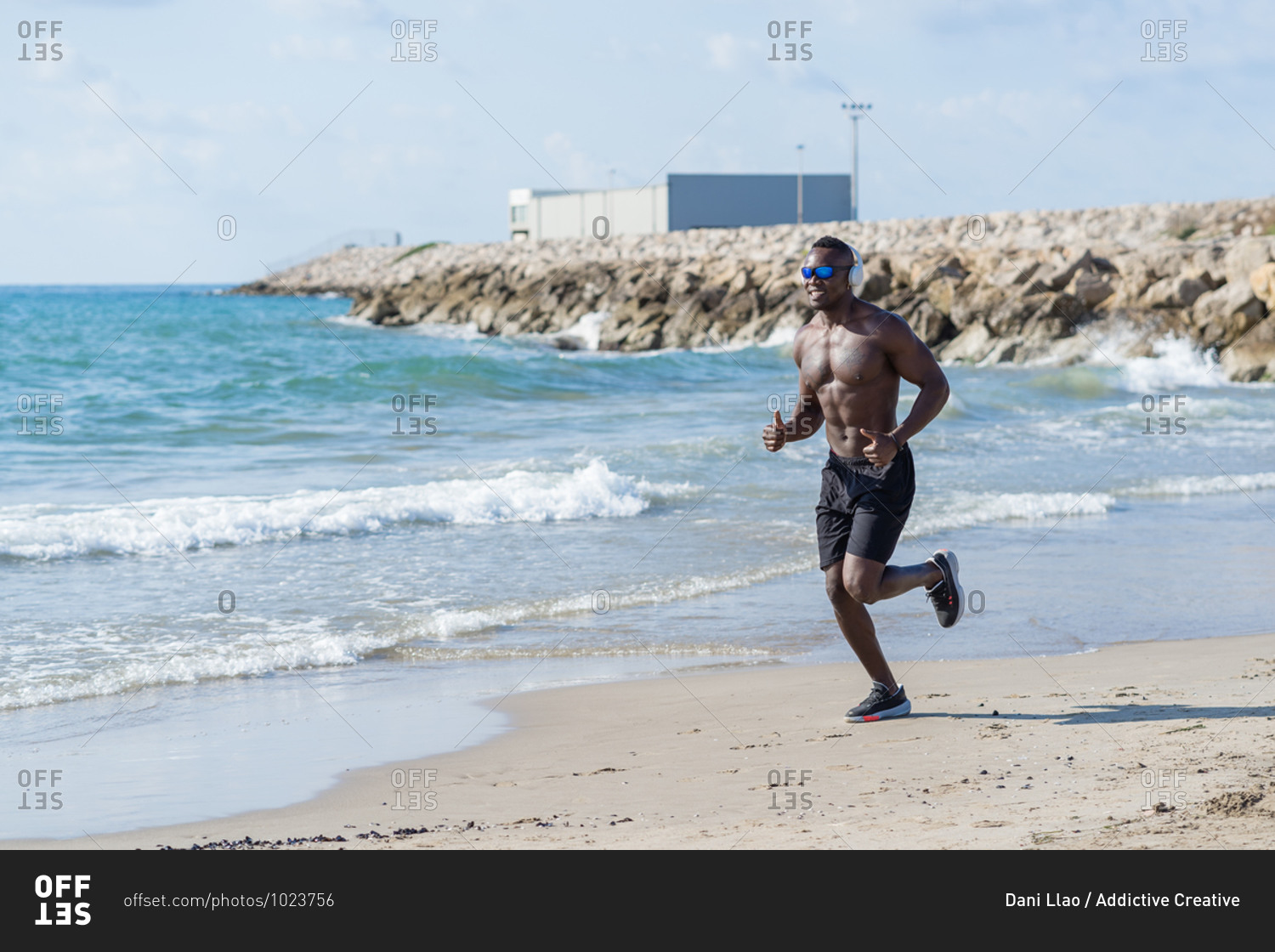 Full body shirtless muscular African American man in headphones and sunglasses jogging on sandy shore near waving sea during workout in sunny day