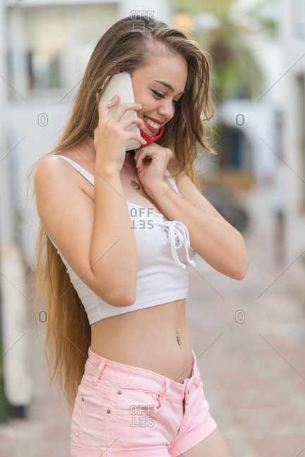 Cheerful female in sexy wear standing on city street and speaking on smartphone while taking off medical mask during COVID 19 and looking away