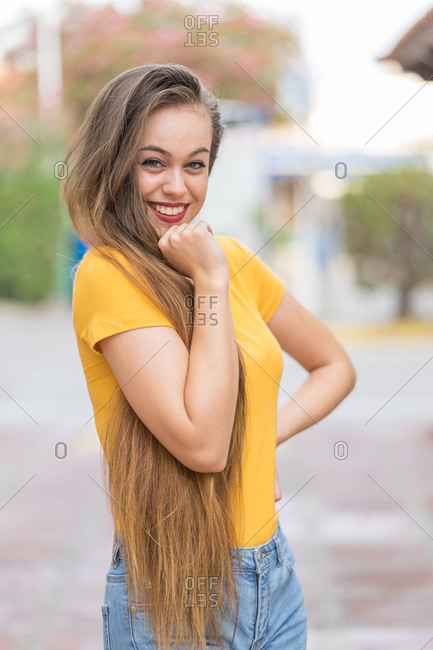 Satisfied female millennial with long hair standing on street and looking at camera while enjoying stroll during weekend