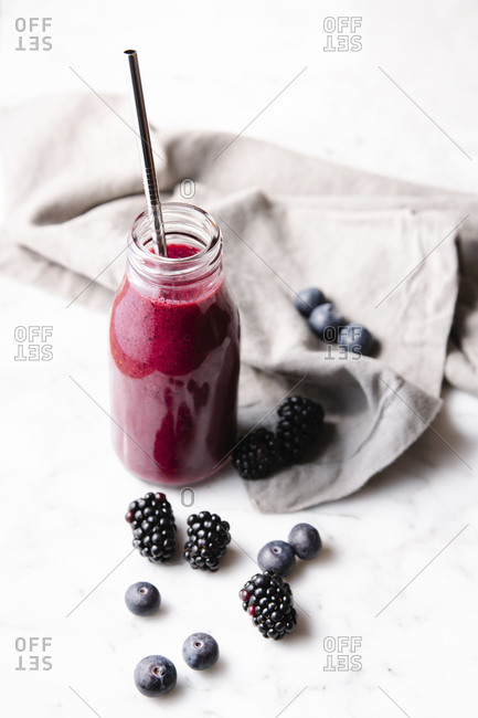 Glass bottle with fresh homemade berry smoothie and a reusable metal straw. Some berries near the bottle on a marble surface. High angle vertical image.