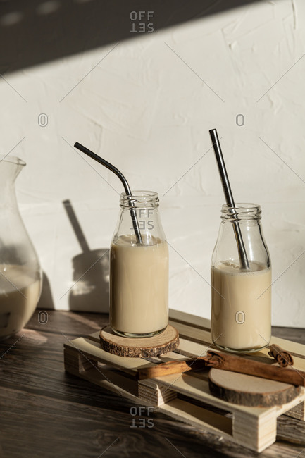 Glass bottles filled with healthy homemade sweet milkshake with cinnamon placed on wooden table against wall with shadow of monstera leaves