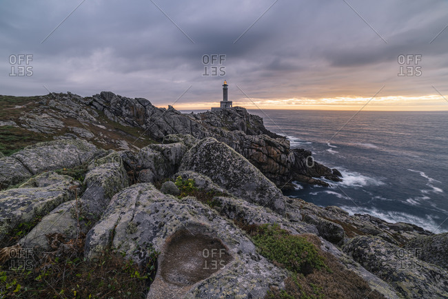 Spectacular scenery of lighthouse located on rough cliff near sea on background of sundown sky