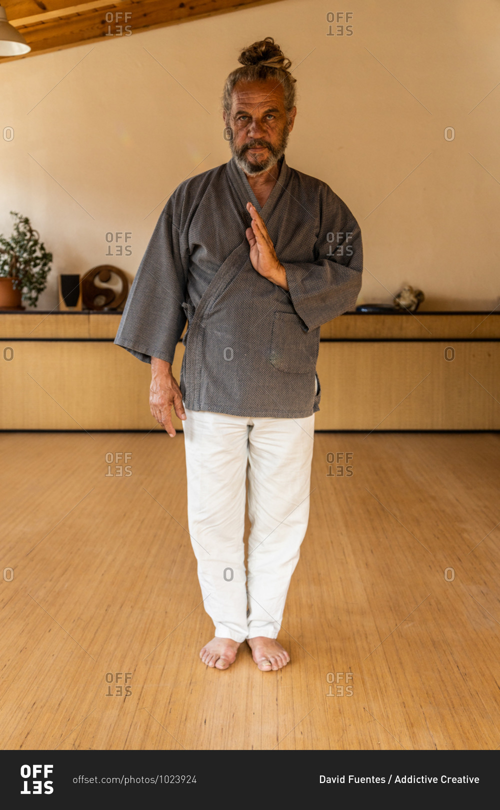Senior bearded ethnic male chi kung practitioner in traditional wear looking at camera while standing in light studio