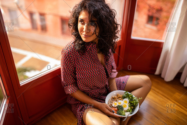 Ethnic latin woman eating a healthy salad on lunch sitting on the floor at home near big windows