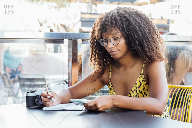 Black self employed woman sitting at table in cafe and working remotely while writing in notepad