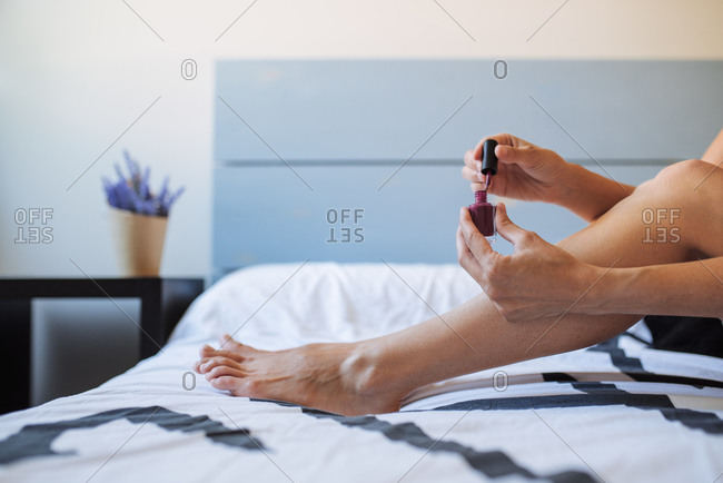 Side view of crop anonymous female sitting on bed and painting nails on toes with bright nail polish during beauty session at home
