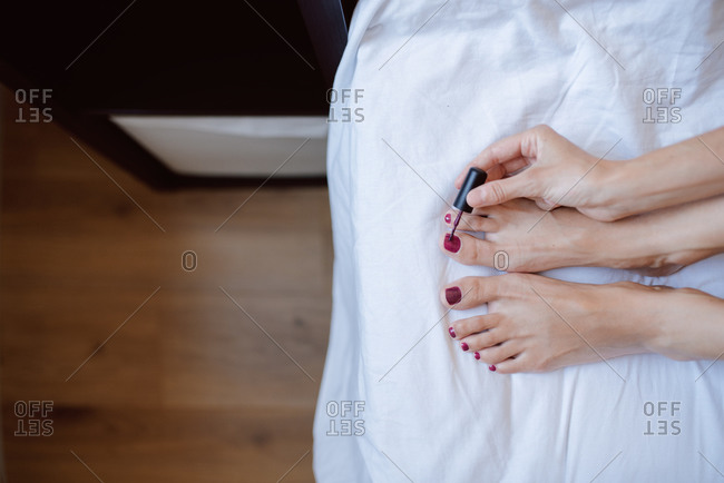 Top view of crop anonymous female sitting on bed and painting nails on toes with bright nail polish during beauty session at home
