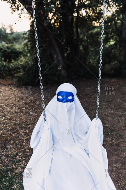 Unrecognizable kid in white ghost costume swinging and playing on playground during Halloween holiday