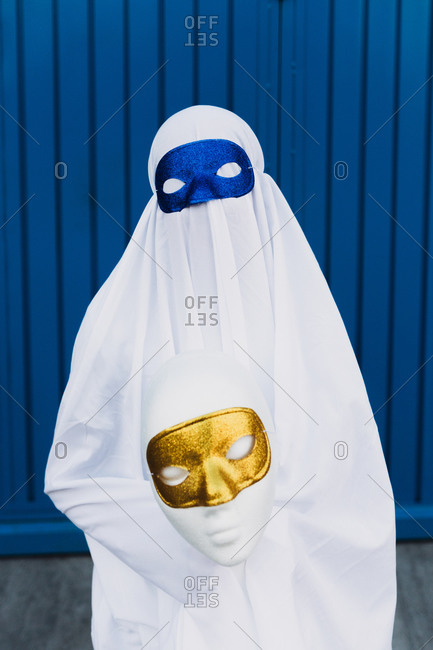 Unrecognizable kid wearing masquerade mask and ghost costume standing in city during Halloween and looking at camera