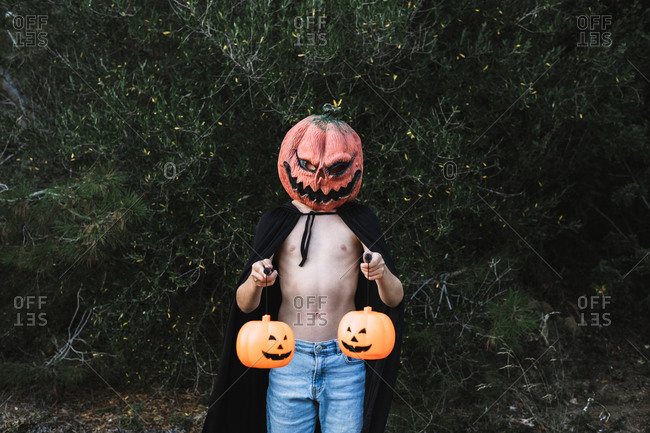 Unrecognizable male wearing creepy mask and black cloak standing with pumpkin lanterns on city street